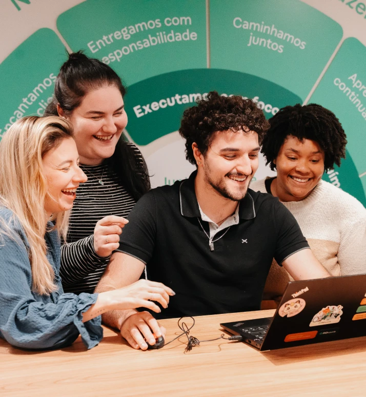 Quatro pessoas lado a lado sorriem em frente a um computador portátil. Da esquerda para a direita, uma mulher branca, de cabelos longos e loiros, usa blusa azul de manga longa. Ao lado dela, uma mulher branca, de cabelos longos castanhos, usa blusa com listras pretas e brancas de manga longa. Na sequência, um homem branco, de cabelos cacheados curtos e castanhos, usa blusa polo preta com detalhes em cinza. Ao lado dele, uma mulher negra de pele clara, com cabelos crespos curtos e castanhos, usa blusa branca e bege de manga longa. O computador portátil está sobre uma mesa e o homem está com a mão sobre o mouse. Ao fundo, parede com itens em tons de verde e palavras em bege.