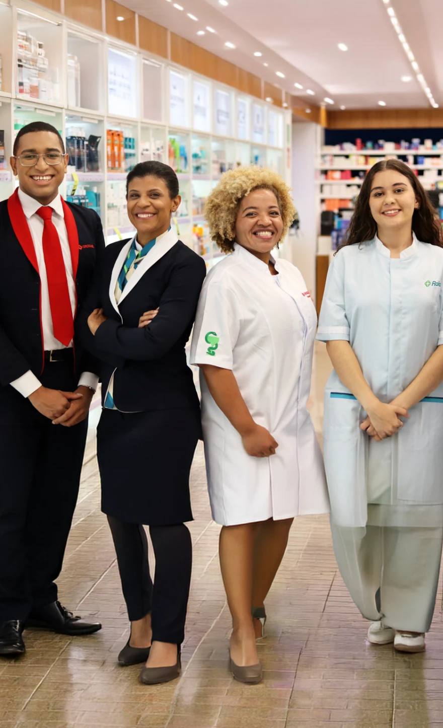 Quatro pessoas lado a lado sorriem. Da esquerda para a direita, um homem jovem, negro, de cabelos castanhos curtos e com óculos de grau. Ele usa camisa branca, gravata vermelha e terno preto com lapela vermelha. Na altura do peito esquerdo do paletó, o logotipo da Drogasil bordado em vermelho. Ao lado dele, uma mulher negra de braços cruzados, cabelos castanhos presos, com um pequeno brinco na orelha esquerda. Ela usa terno preto com lapela branca e lenço com listras bege e em tons de verde e azul no pescoço. Na sequência, uma mulher negra de cabelos crespos, curtos e loiros. Ela usa jaleco branco com bordado de Taça de Hígia em tons de verde no braço direito. Na altura do peito esquerdo, o logotipo da Drogasil bordado em vermelho. Ao lado dela, uma mulher jovem, branca de cabelos castanhos longos, com pequenos brincos na orelha esquerda e piercings no nariz. Ela usa jaleco branco com o logotipo da Raia bordado na altura do peito esquerdo. Ao fundo e desfocado, diversas prateleiras iluminadas com luz branca, com medicamentos e itens de cuidado pessoal. Um pequeno grafismo de linha fina e verde forma um retângulo em volta do busto das quatro pessoas. Abaixo do grafismo, o nome do relatório dentro de um retângulo laranja, o ano do relatório dentro de um quadrado verde-claro e o logotipo da R D Saúde dentro de um retângulo verde-escuro. As três formas possuem bordas arredondadas. O logotipo da R D Saúde é composto por dois elementos, sendo uma cruz com centro arredondado, cercada por três triângulos arredondados de cor verde-clara, seguida das letras R D maiúsculas e Saúde em letras minúsculas, todas em branco.